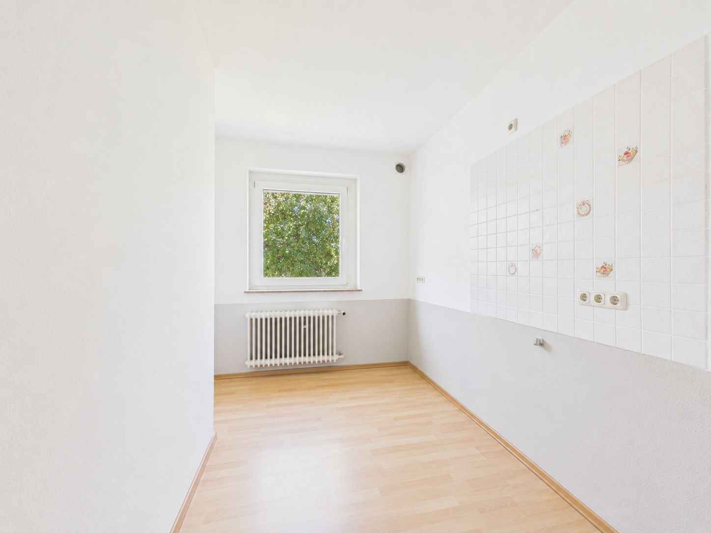 Heller leerer Raum mit Fenster, Heizkörper und teils gefliester Wand, heller Holzboden.