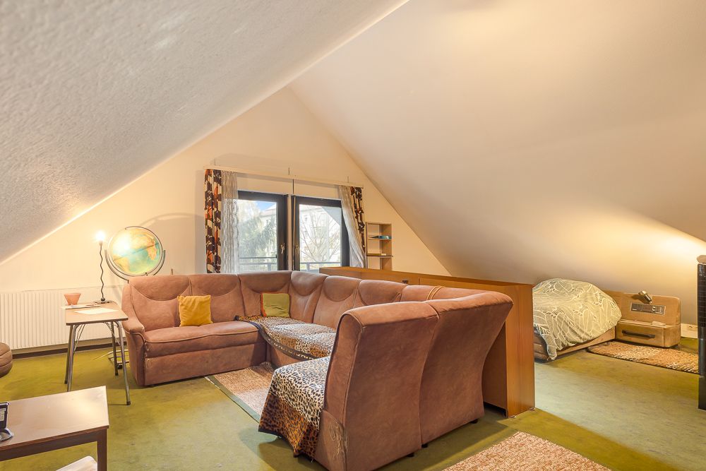 Wohnzimmer im Dachgeschoss mit Ecksofa, Teppichen und Bett unter schrägem Dach.