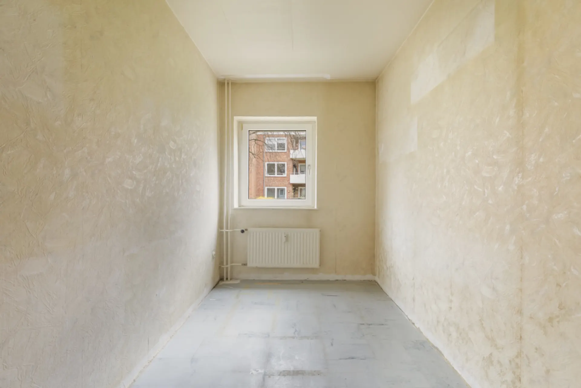 Leerer, schmaler Raum mit einem Fenster und Heizkörper an der Stirnwand.
