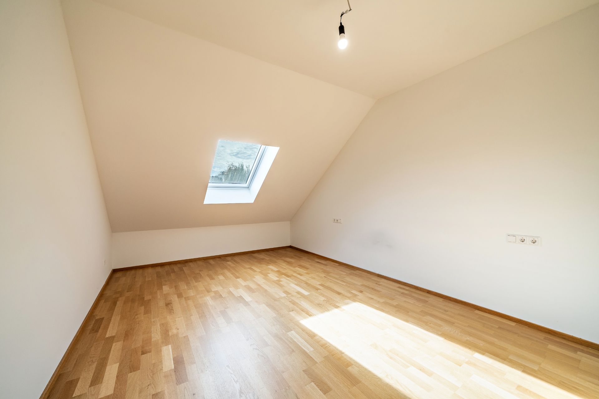 Leeres Zimmer mit schräger Decke, Dachfenster und hellem Holzboden.