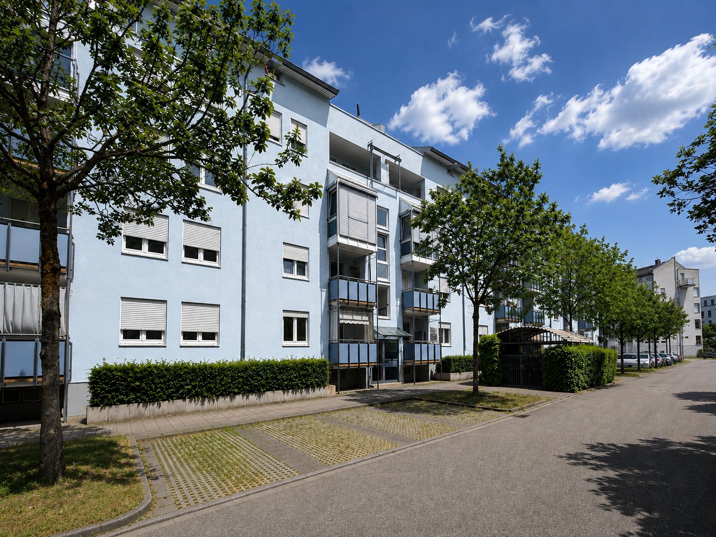 Mehrfamilienhaus mit Balkonen, Bäumen und Parkplätzen an einer ruhigen Straße.