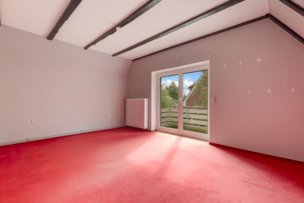 Leeres Zimmer mit rotem Teppich, weißen Wänden und Balkontür, sichtbare Deckenbalken.