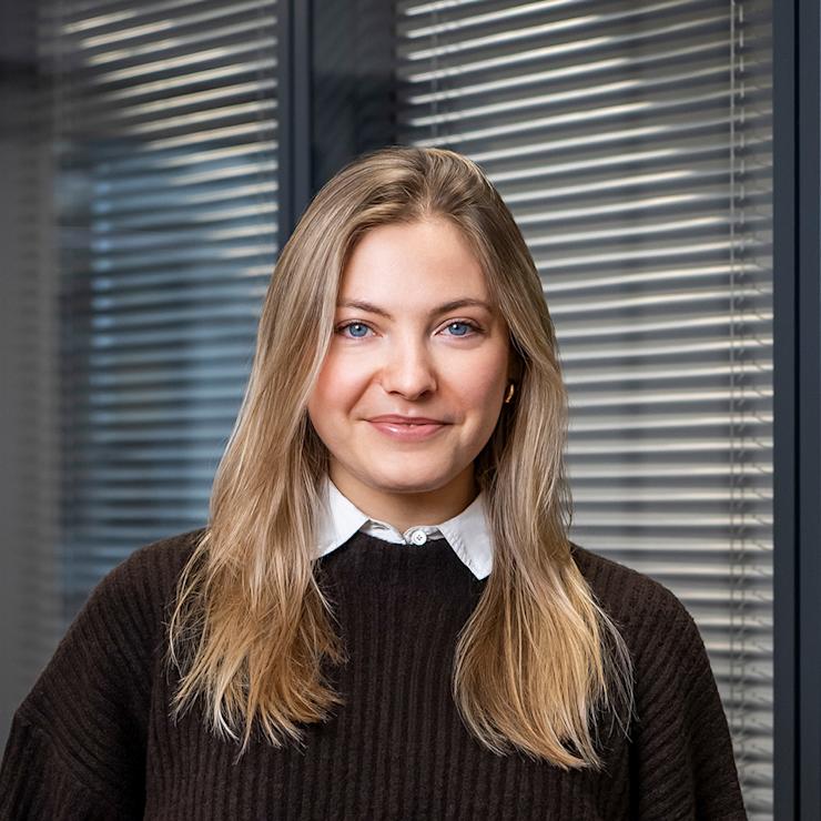 Junge Frau mit langen blonden Haaren steht vor Fenster mit Jalousien.