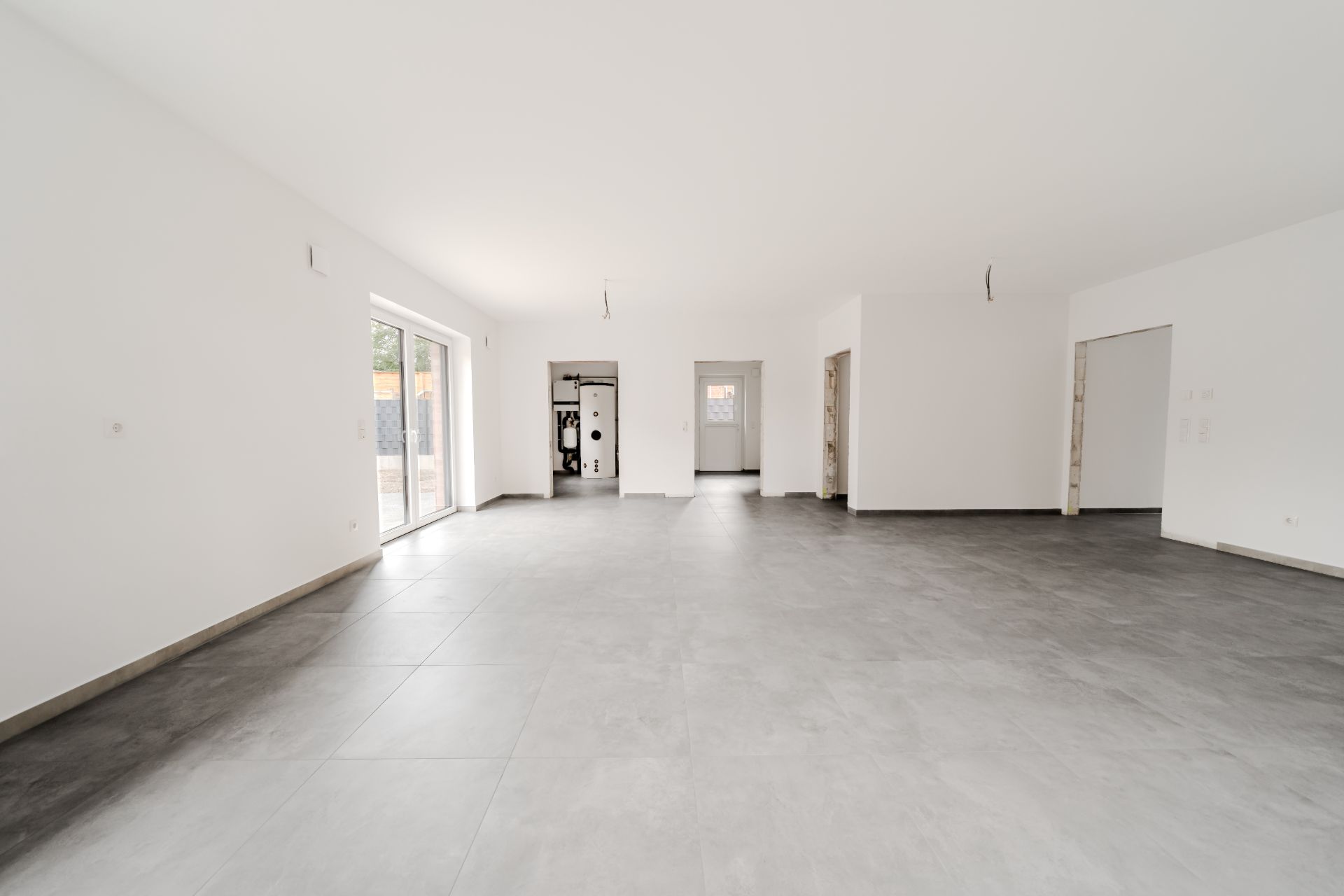Leerer, heller Raum mit grauem Fliesenboden und weißen Wänden, große Fenster links.