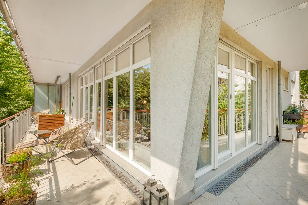 Großer Balkon mit Sitzmöbeln, Pflanzen und großen Fenstern zu einem Wohngebäude.