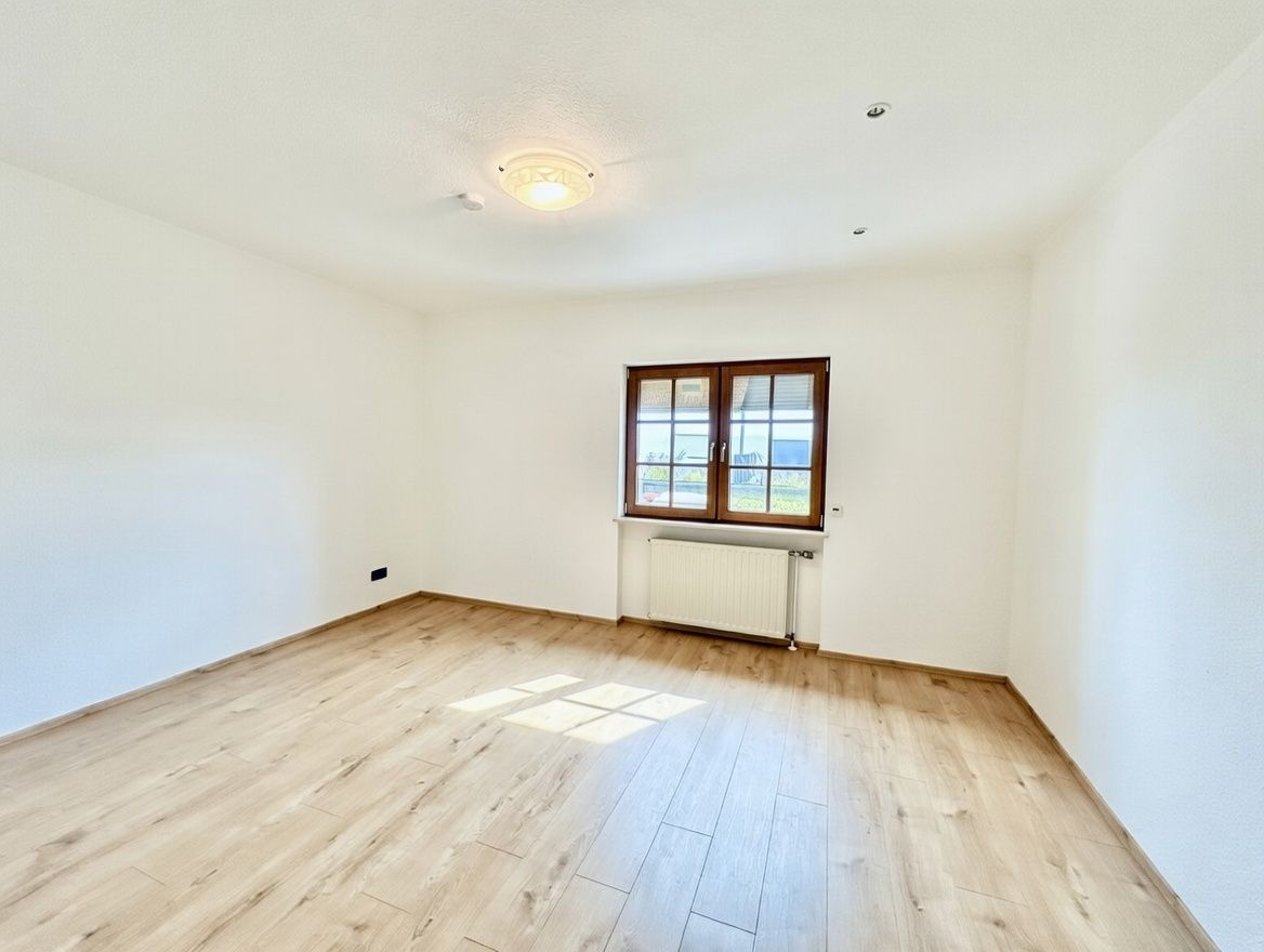 Leerer, heller Raum mit Holzfußboden, weiß gestrichenen Wänden und einem Fenster mit Heizkörper darunter.