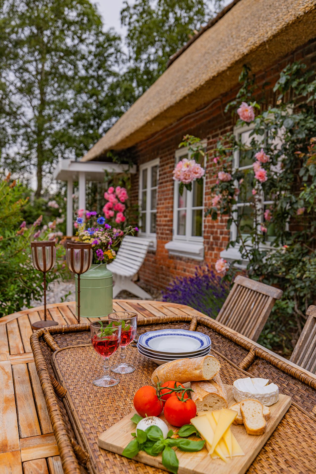 Holztisch im Garten mit Käse, Brot, Tomaten und zwei Gläsern Getränk vor einem Backsteinhaus mit Blumen.