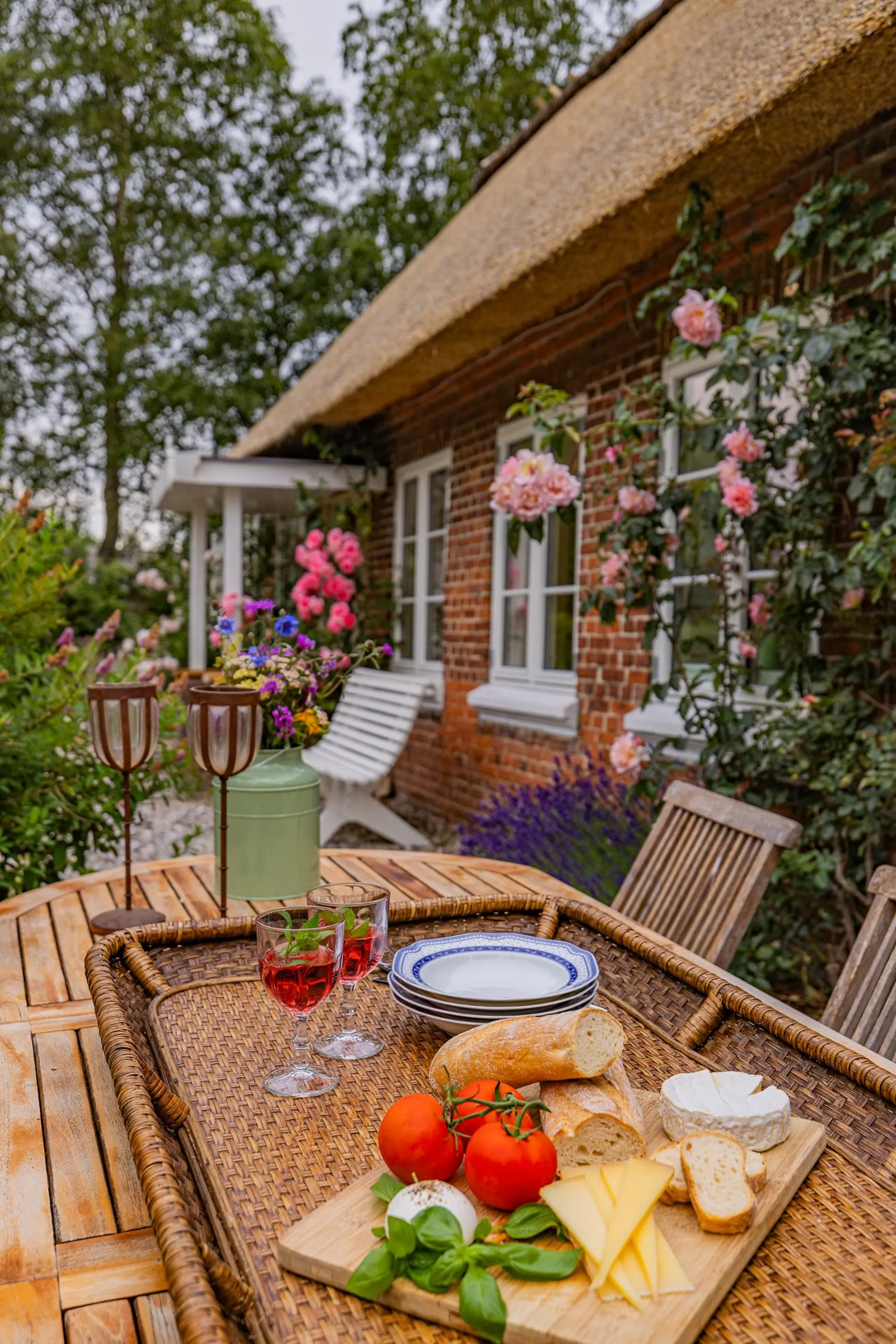 Holztisch im Garten mit Käse, Brot, Tomaten und zwei Gläsern Getränk vor einem Backsteinhaus mit Blumen.