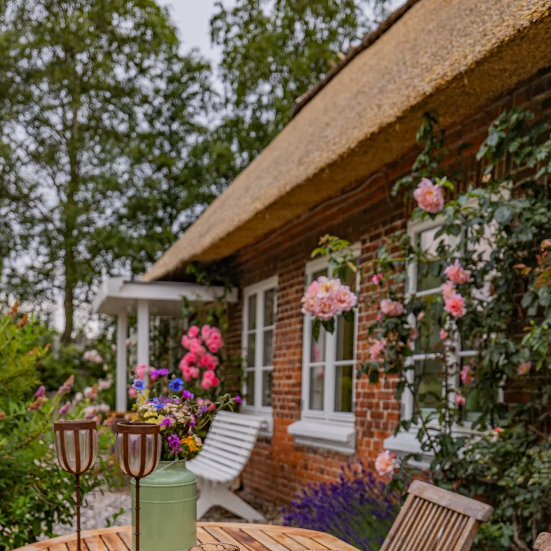 Holztisch im Garten mit Käse, Brot, Tomaten und zwei Gläsern Getränk vor einem Backsteinhaus mit Blumen.