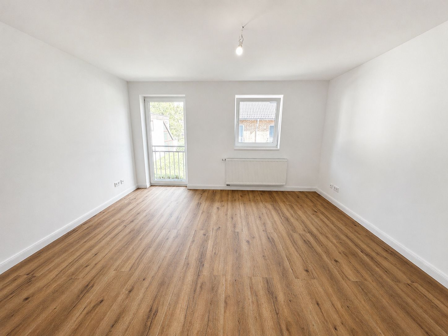 Helles, leeres Zimmer mit Holzfußboden, Fenster und Tür zum Balkon.