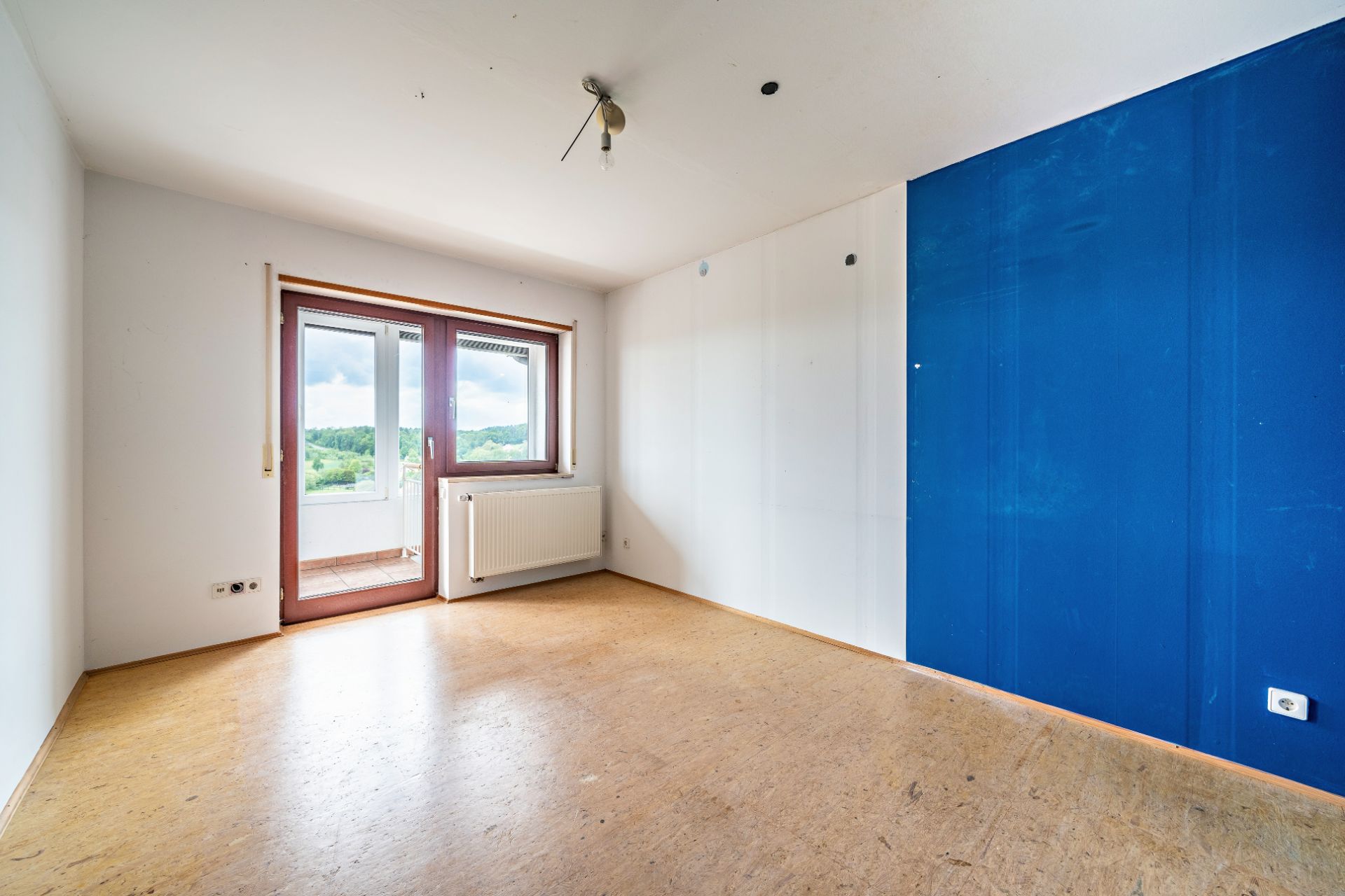 Leeres Zimmer mit Holzboden, blauer Wand und Fenster mit Zugang zum Balkon.