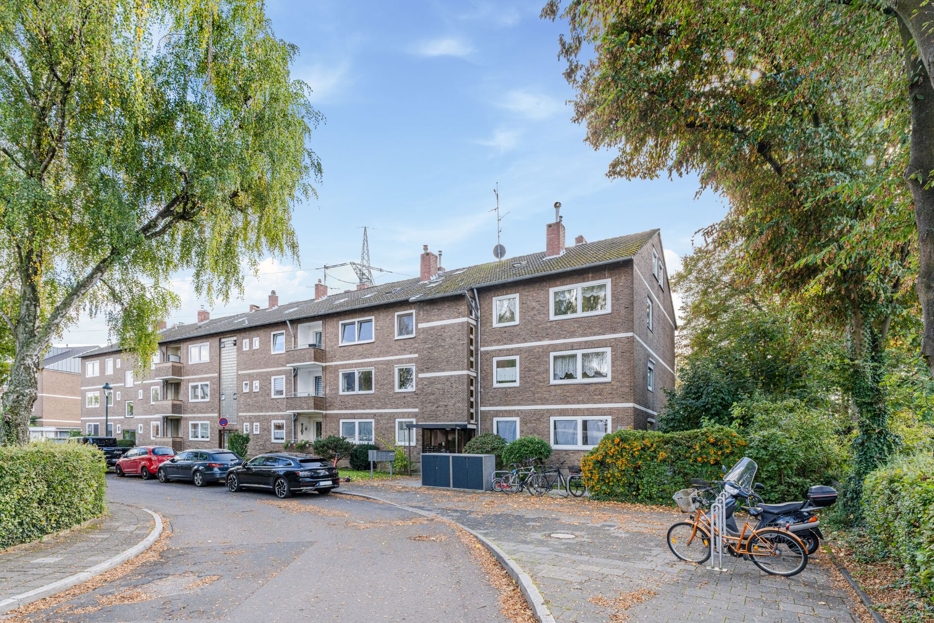 Mehrfamilienhaus aus Backstein mit geparkten Autos, Fahrrädern und Bäumen am Straßenrand.