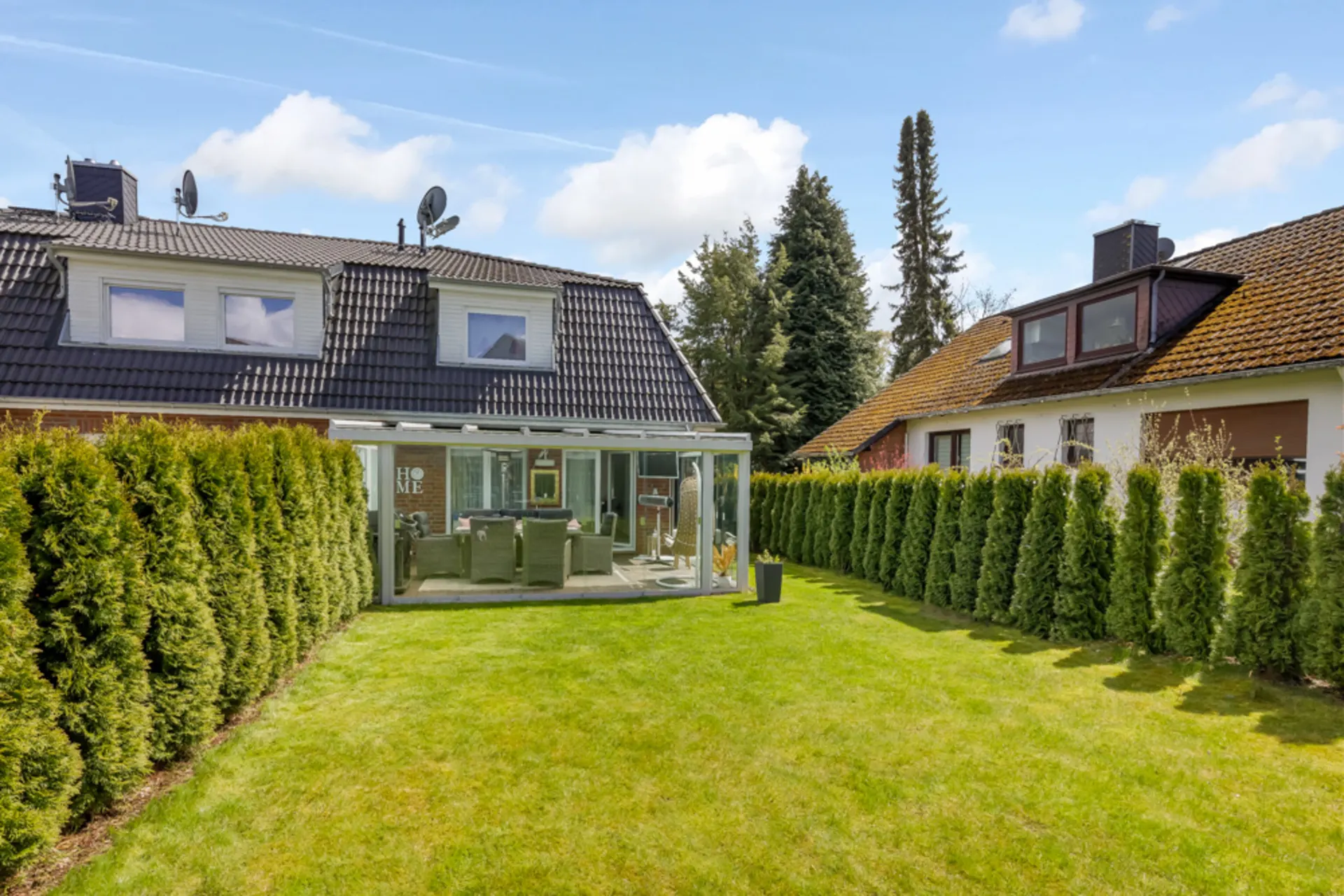 Garten mit Rasen, Terrasse mit Glasüberdachung und zwei Wohnhäusern im Hintergrund.