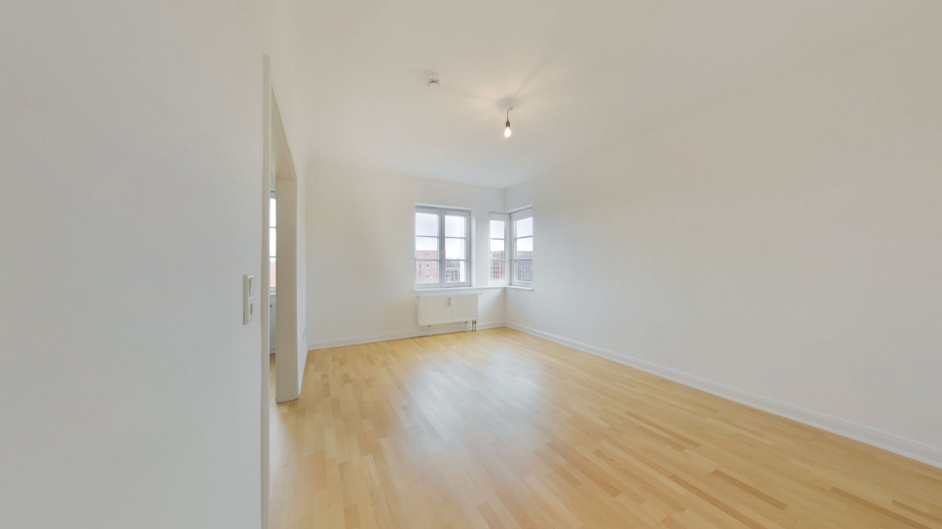 Leeres, helles Zimmer mit Holzfußboden und drei Fenstern.