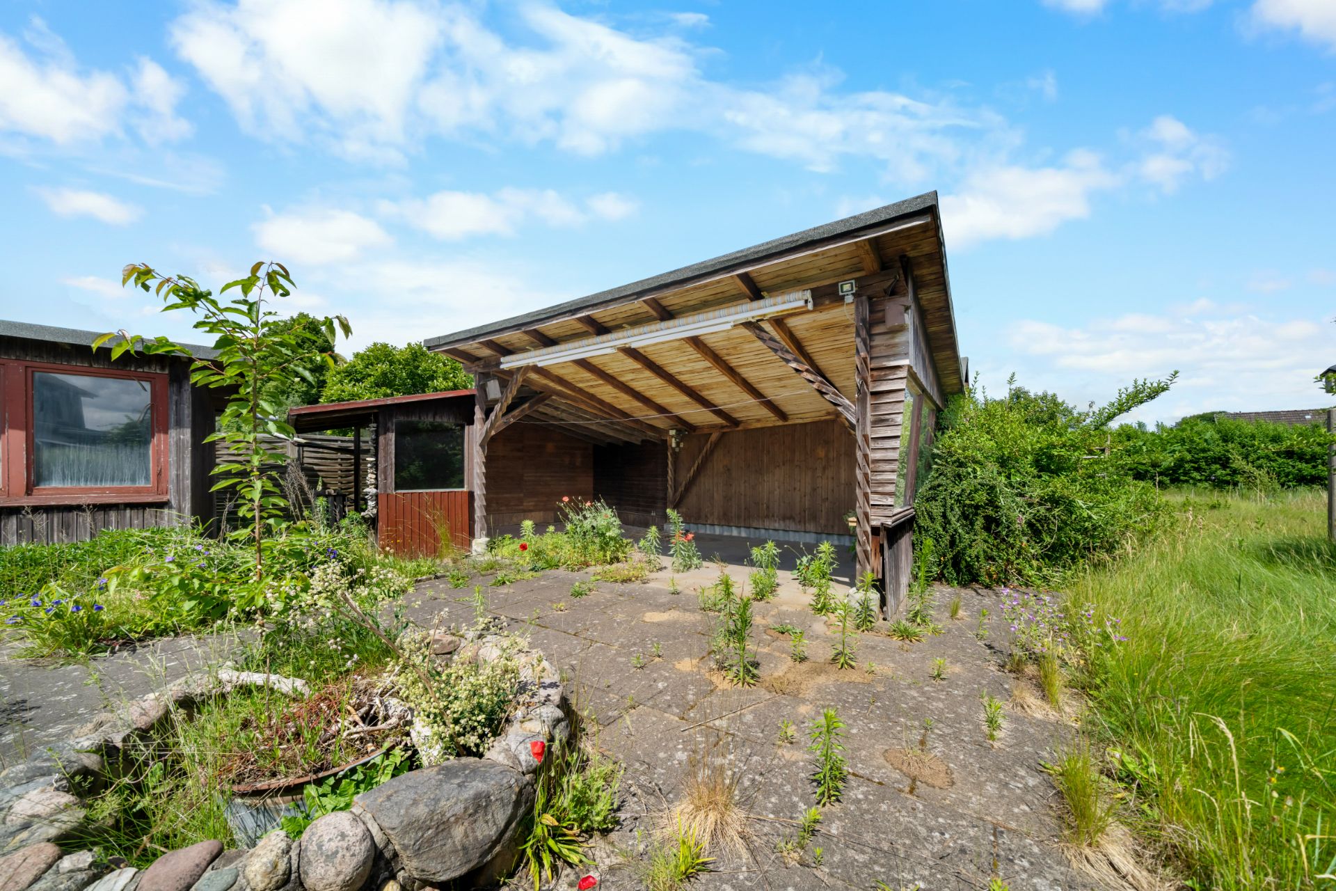 Holzüberdachung mit gepflastertem Hof, wilden Pflanzen und kleinem Gartenbereich.