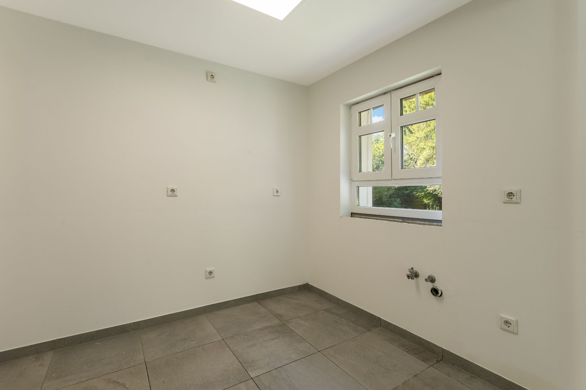 Leerer Raum mit hellen Wänden, Fliesenboden und Fenster, mehrere Steckdosen sichtbar.