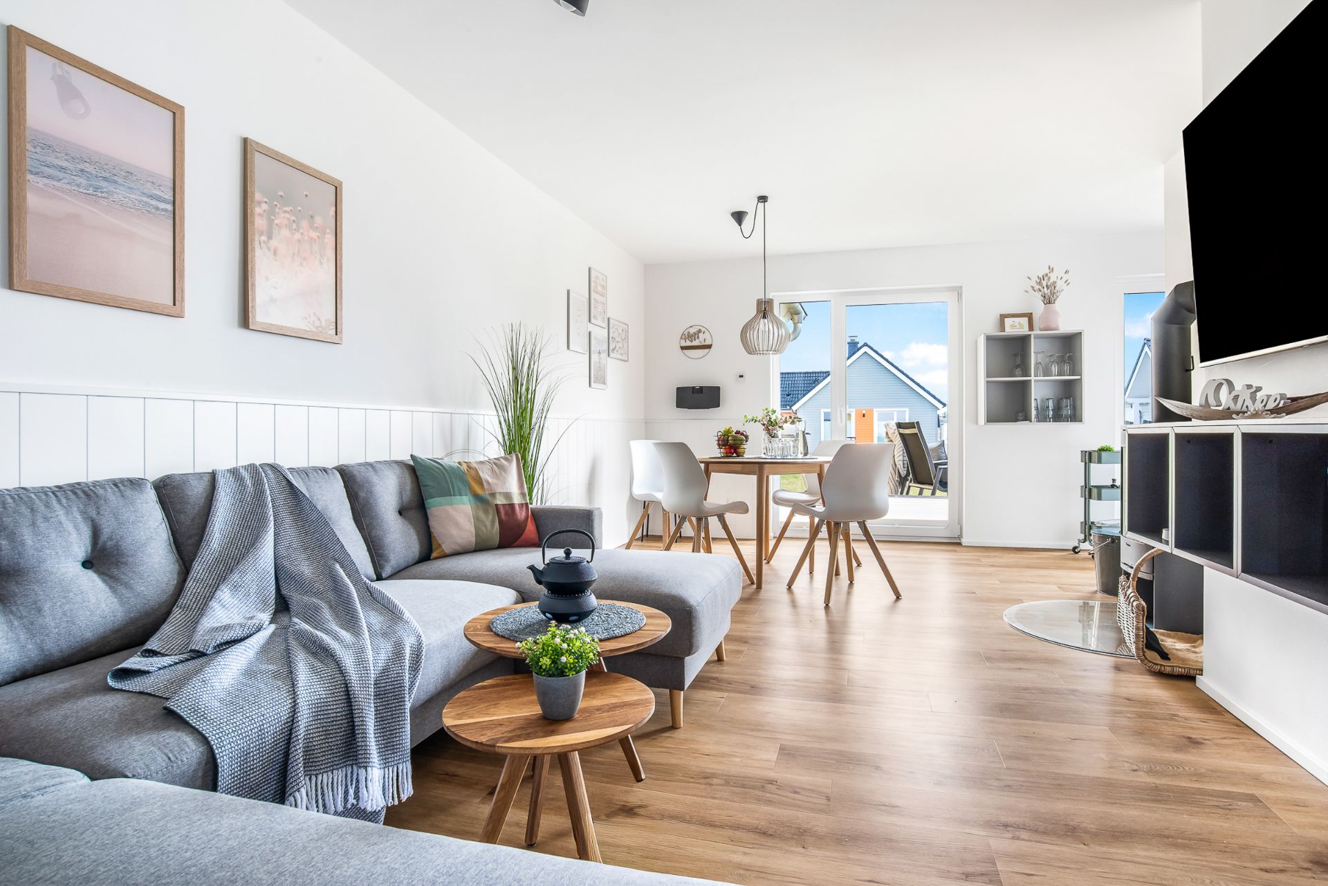 Helles Wohnzimmer mit grauem Sofa, Essbereich, Holzfußboden und großen Fenstern.