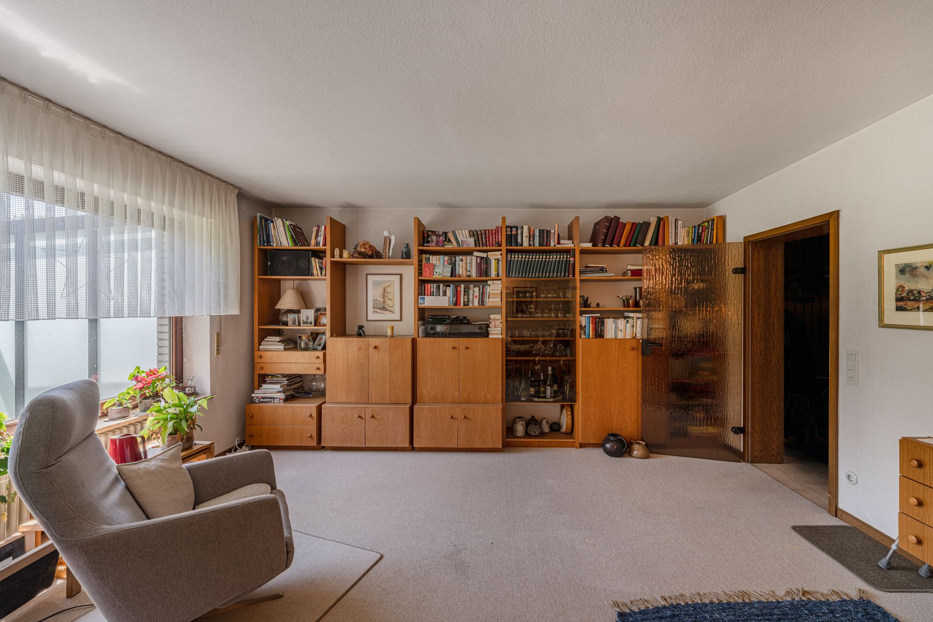 Wohnzimmer mit grauem Sessel, Regalwand aus Holz und großen Fenstern mit weißen Vorhängen.