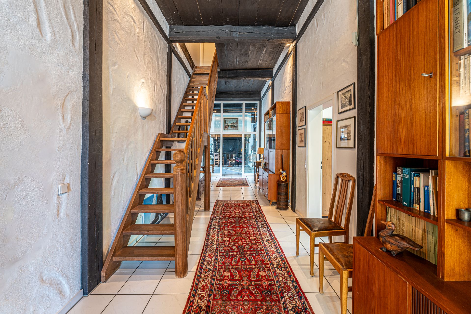 Flur mit roten Teppichläufer, Holztreppe, Bücherregal und zwei Holzstühlen.