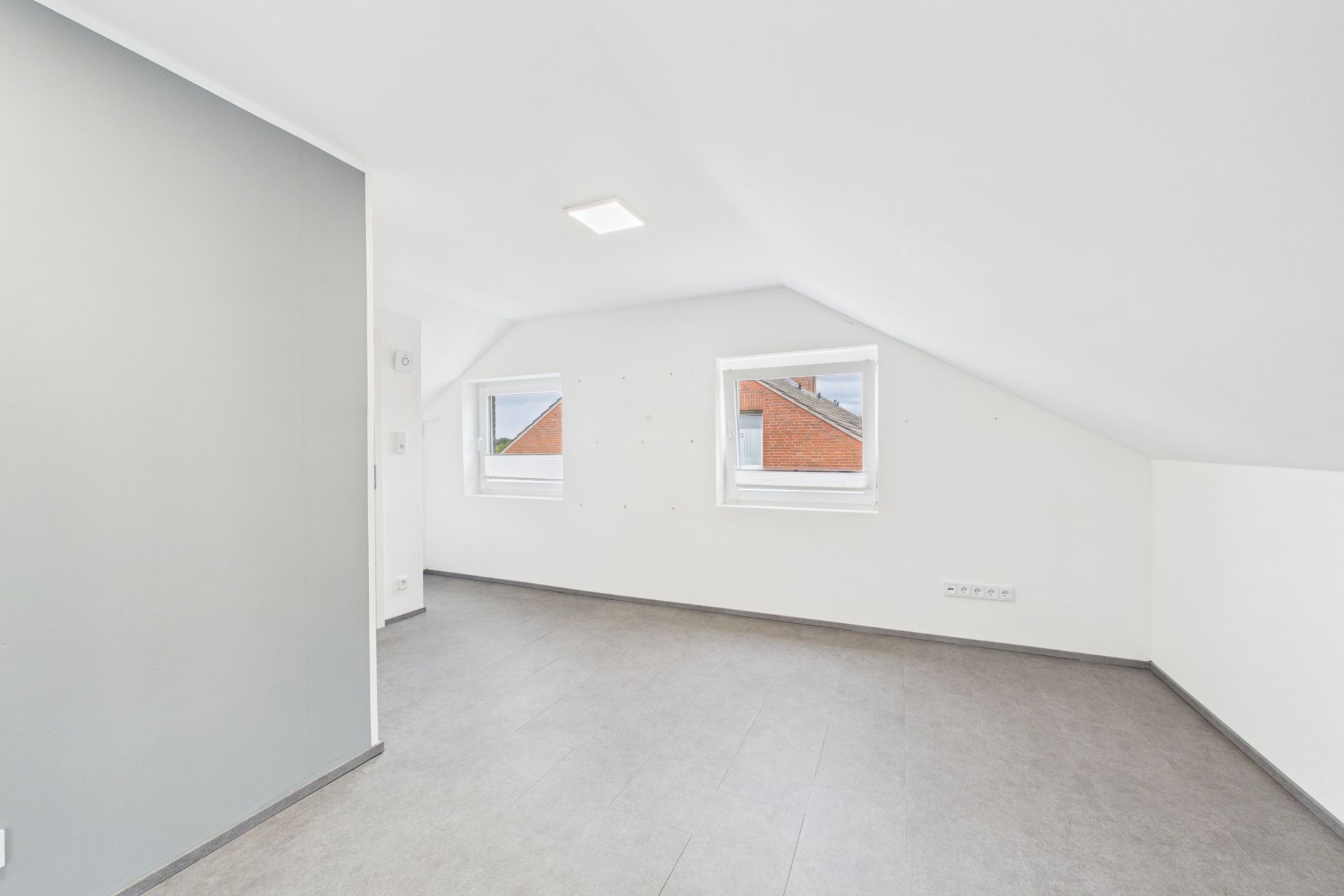 Heller Dachgeschossraum mit zwei Fenstern und schräger Decke, grauer Boden, weiße Wände.