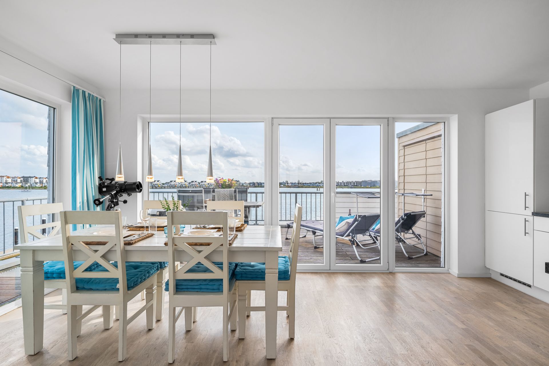 Essbereich mit weißen Stühlen, Tisch, blauen Kissen und Seeblick durch große Fenster und Balkon.