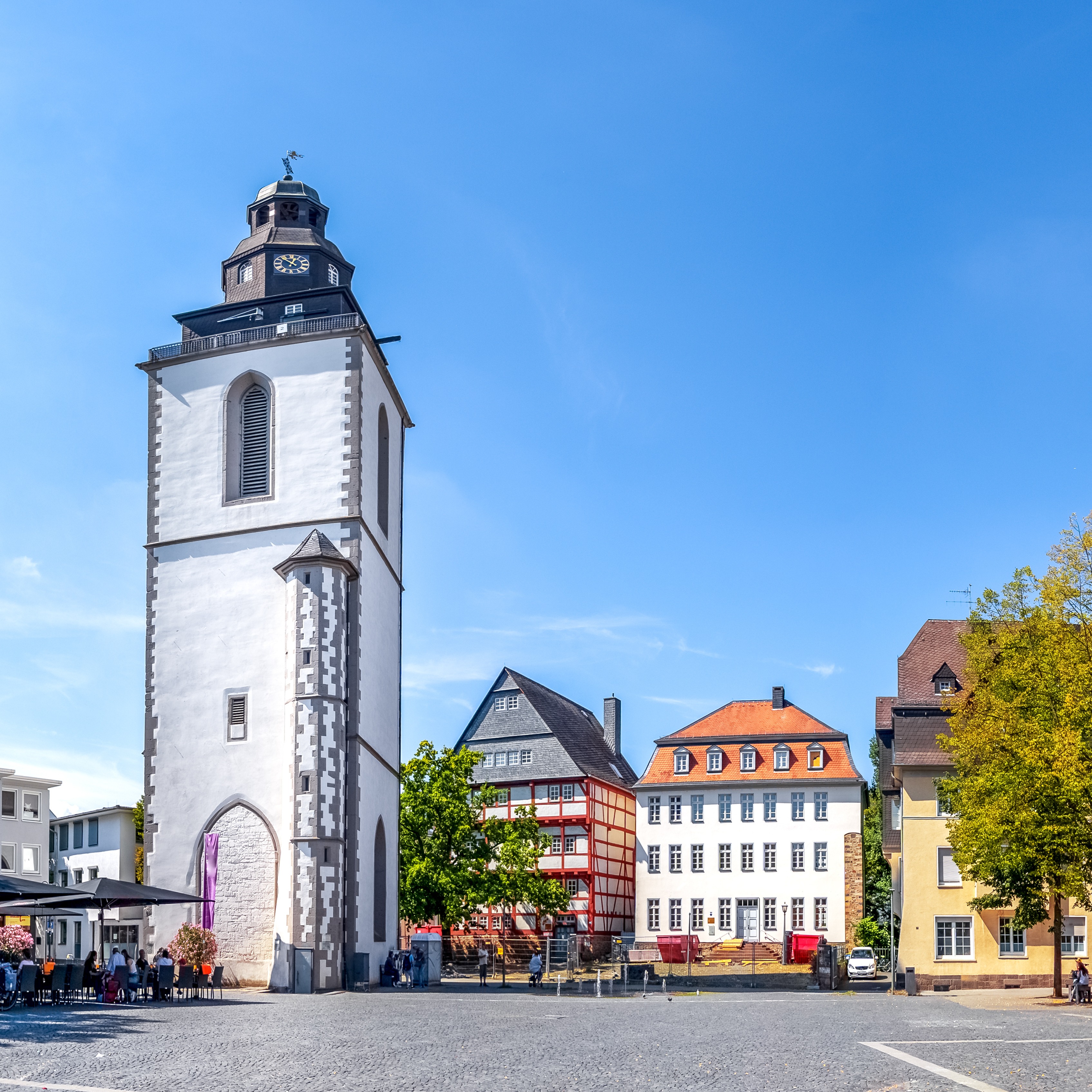 Weißer Kirchturm auf einem sonnigen Platz, umgeben von Bäumen und Wohnhäusern.