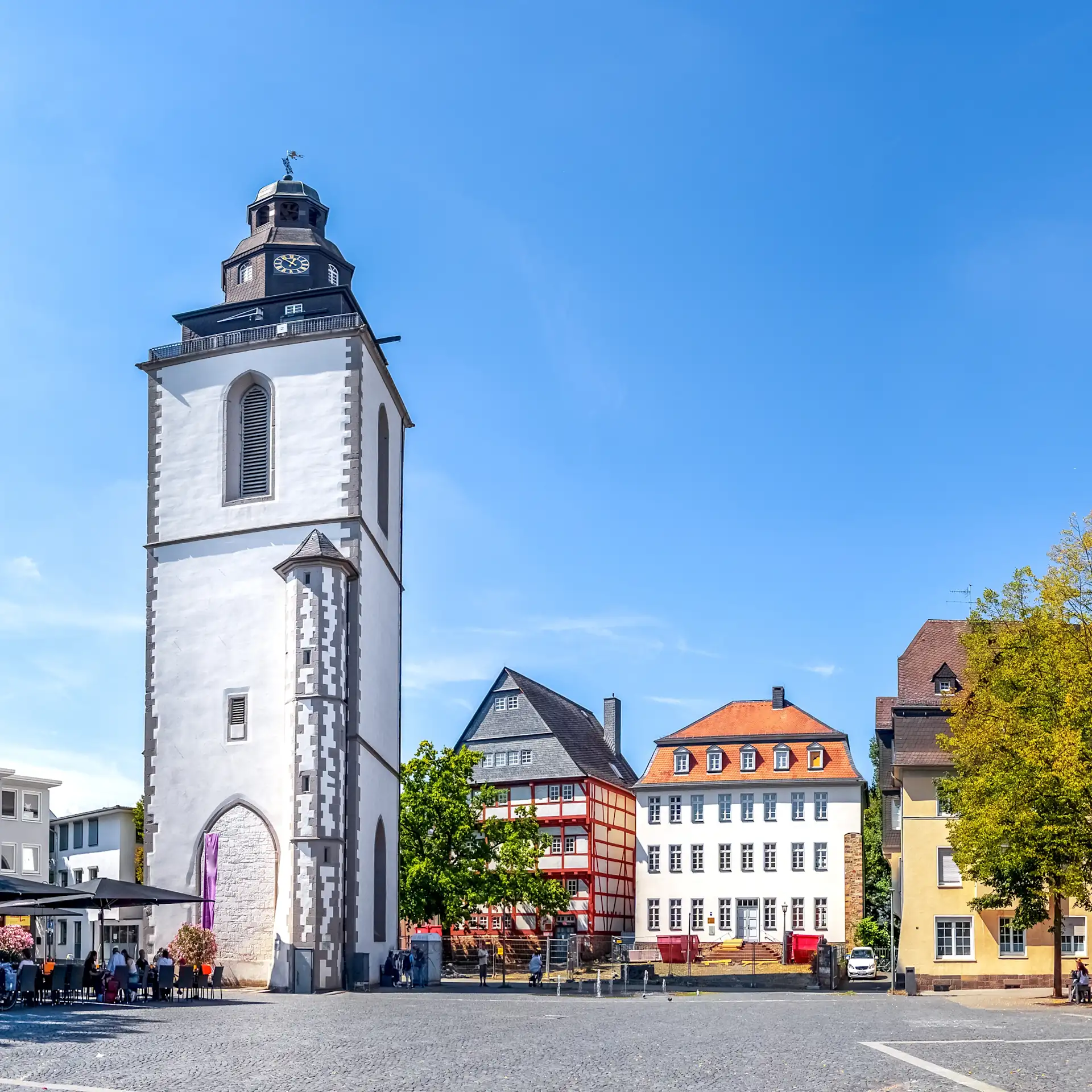 Weißer Kirchturm auf einem sonnigen Platz, umgeben von Bäumen und Wohnhäusern.