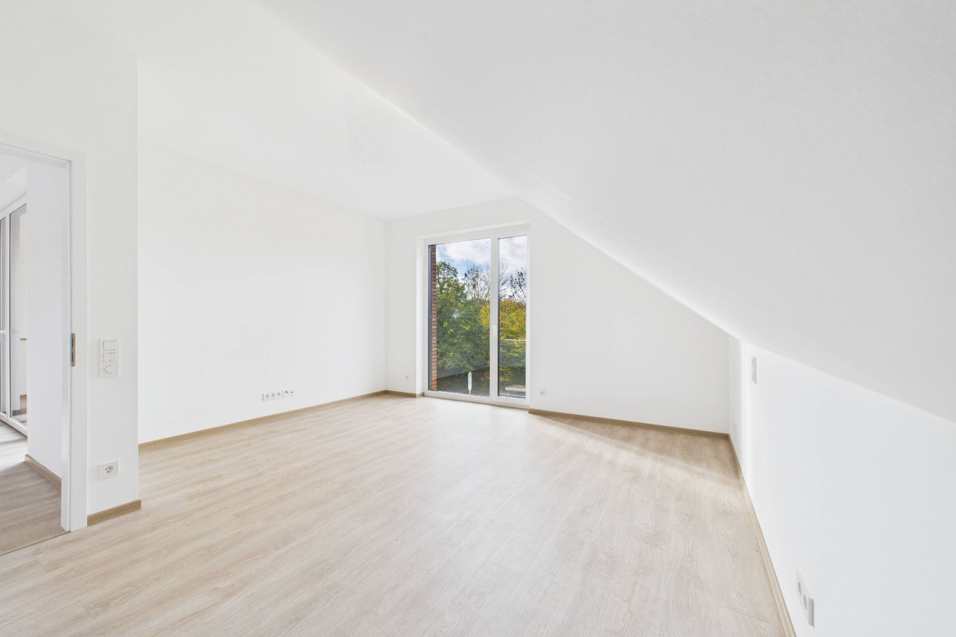 Helles, leeres Zimmer mit Holzfußboden, Dachschräge und großem Fenster mit Blick ins Grüne.