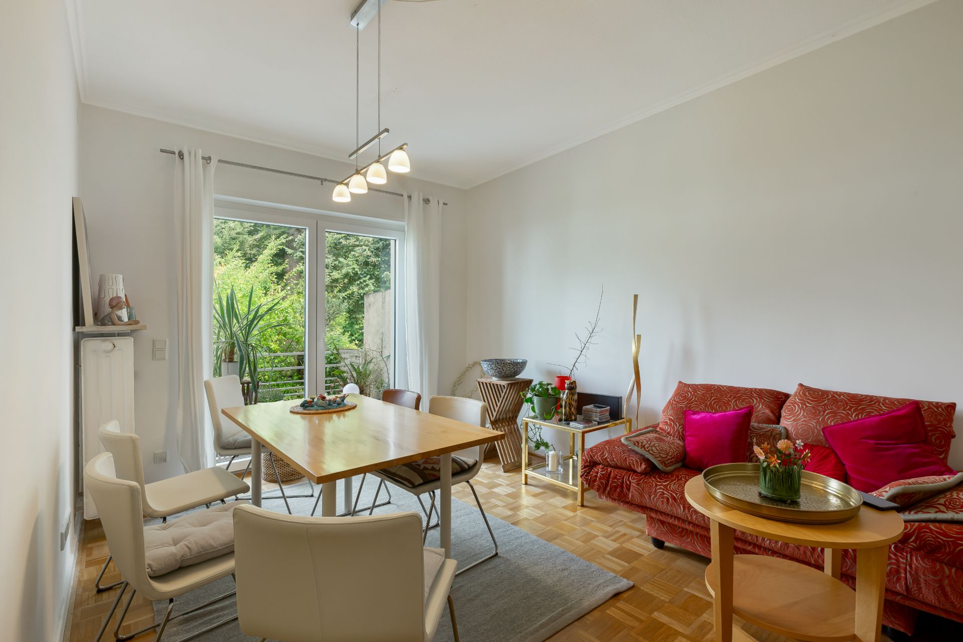 Helles Wohnzimmer mit Esstisch, Stühlen, rotem Sofa und Glas­tür zum Balkon mit Blick ins Grüne.