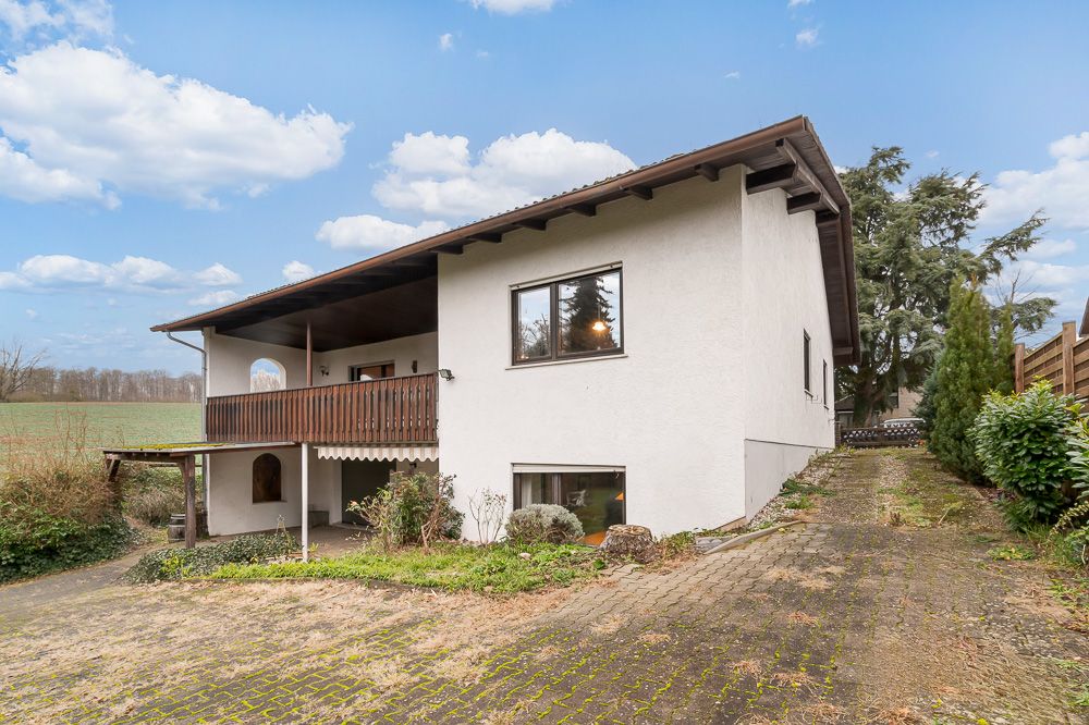 Zweistöckiges Haus mit weißer Fassade, Balkon und gepflasterter Auffahrt, umgeben von Natur.