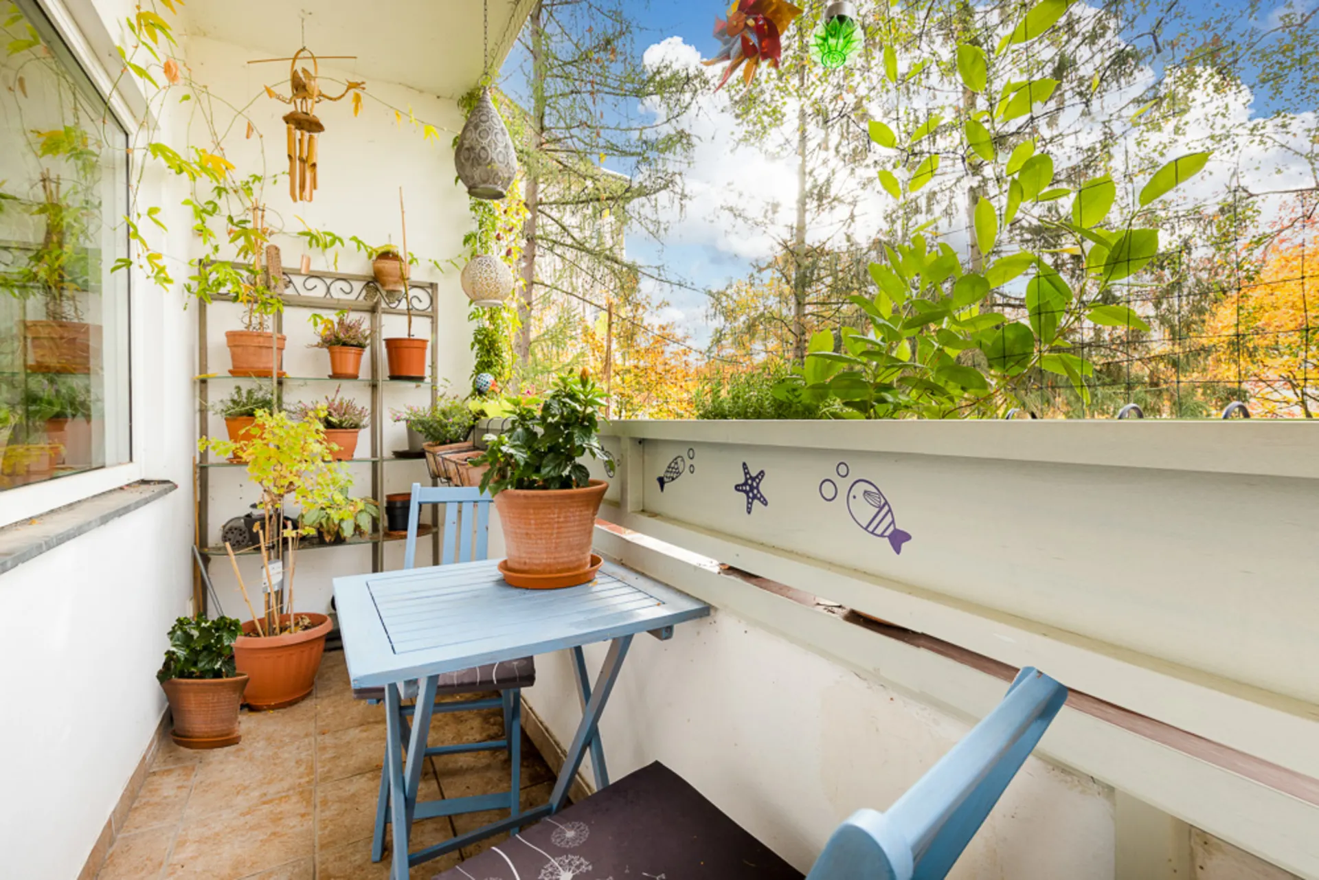 Kleiner Balkon mit blauem Tisch, zwei Stühlen und vielen Pflanzen in Töpfen.