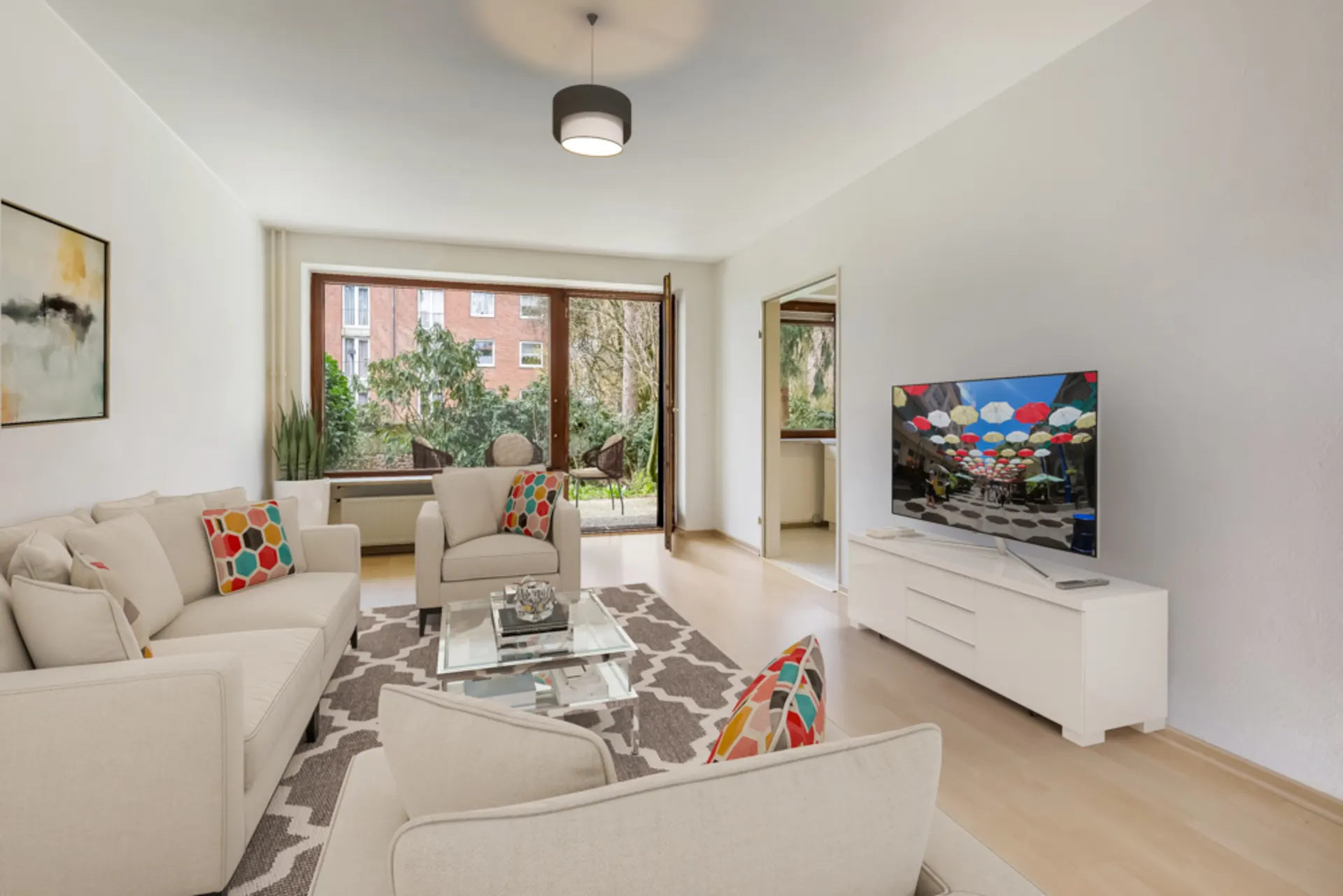 Helles Wohnzimmer mit beigen Sofas, bunten Kissen, TV und großem Fenster zum Garten.