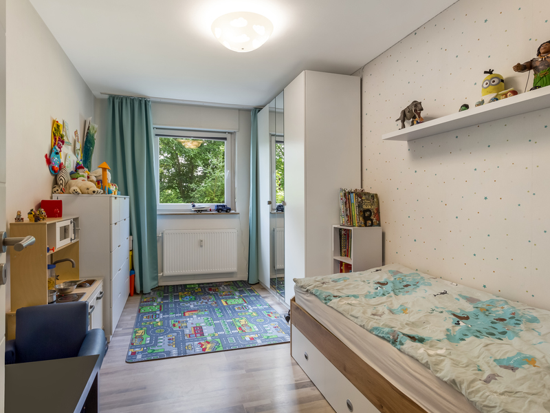 Helles Kinderzimmer mit Bett, Regal, buntem Teppich und Fenster mit grünen Vorhängen.