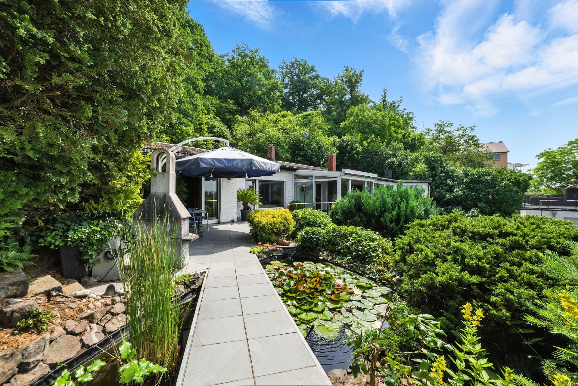 Moderner Garten mit Teich, Terrasse, Sonnenschirm und vielen grünen Pflanzen vor einem weißen Haus.