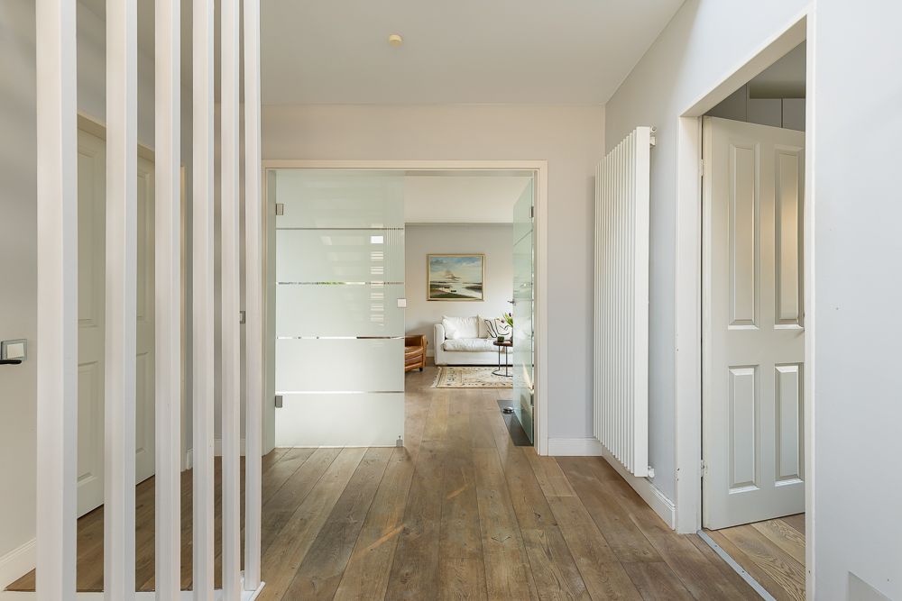 Heller Flur mit Holzboden, Glas­tür und Blick in ein Wohnzimmer mit Sofa und Bild an der Wand.