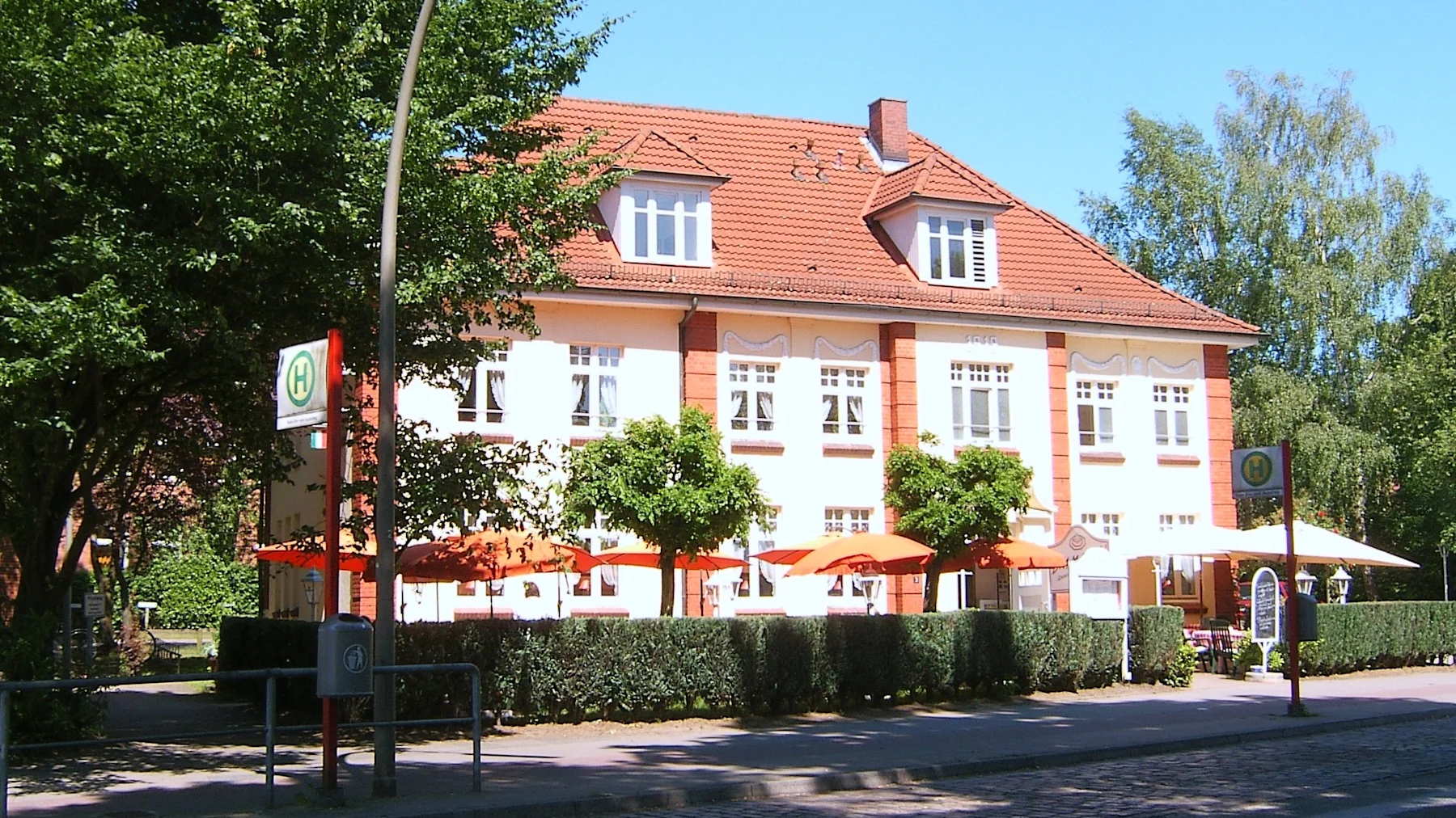 Großes Wohnhaus mit rotem Dach, umgeben von Bäumen und Sonnenschirmen vor dem Gebäude.