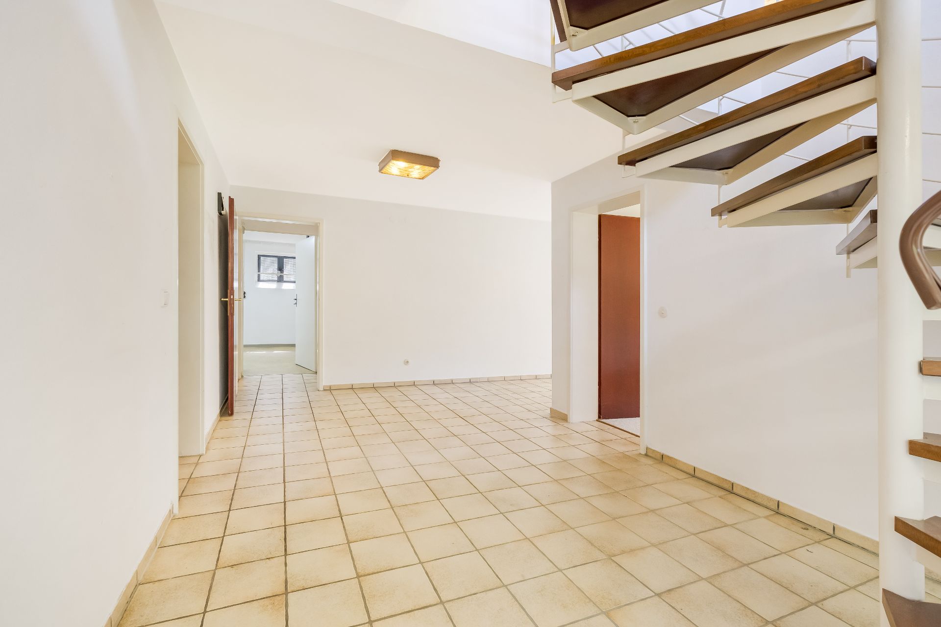 Heller Flur mit Fliesenboden, weißer Wand und moderner Treppe aus Holz und Metall.