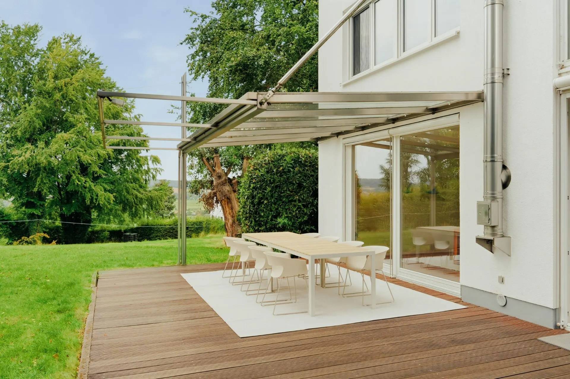 Holzterrasse mit Essbereich, Tisch und Stühlen neben einem weißen Wohnhaus, Glasüberdachung, Garten im Hintergrund.