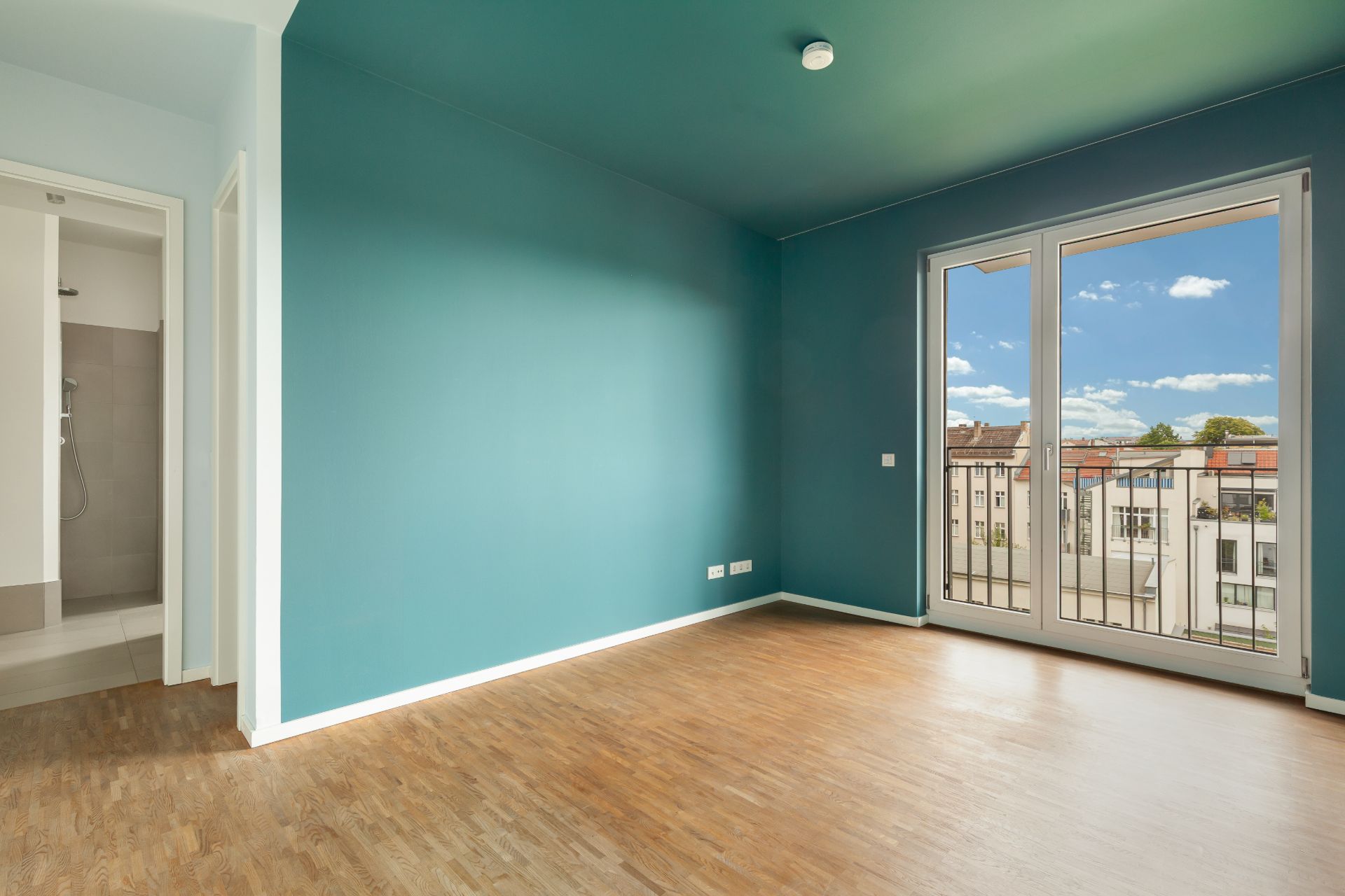 Leeres Zimmer mit Holzboden, türkisfarbigen Wänden und großen Fenstern mit Blick nach draußen.