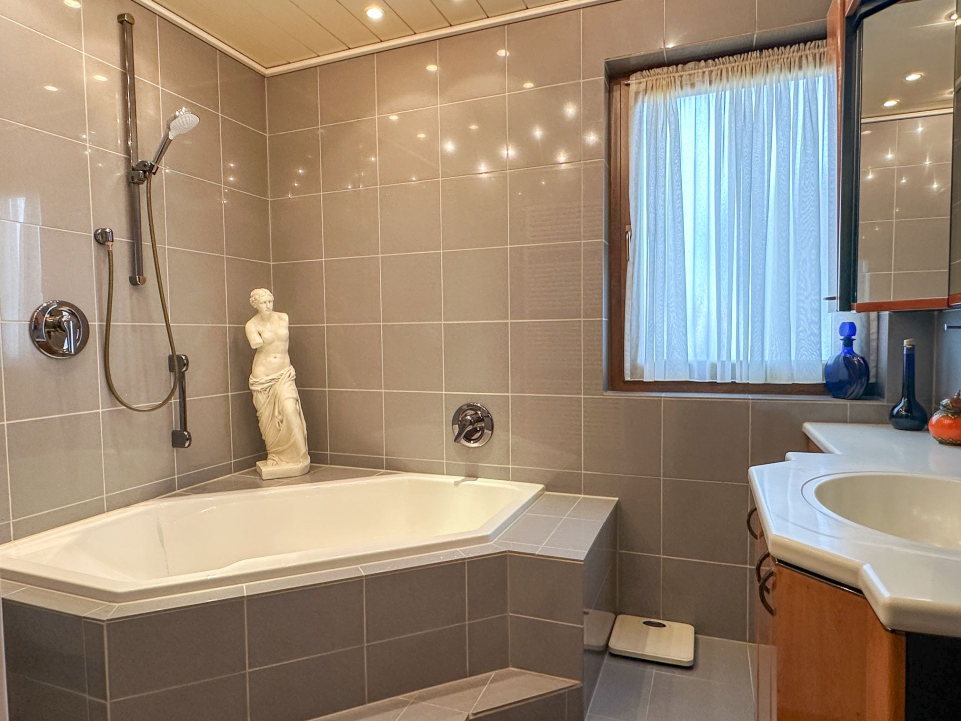Modernes Badezimmer mit Eckbadewanne, Statue, Waschbecken, Fenster mit Vorhang und Handbrause.