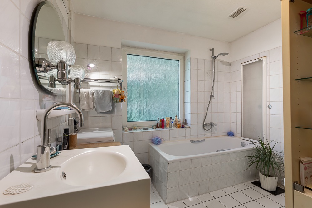 Helles Badezimmer mit Badewanne, Waschbecken, Spiegel und Fenster. Pflanze steht neben der Badewanne.