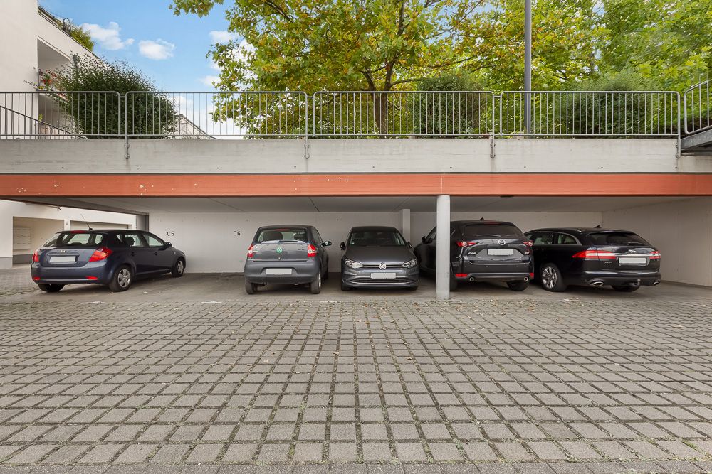Überdachte Parkplätze mit fünf Autos in einer Wohnanlage, Baum und Geländer im Hintergrund.