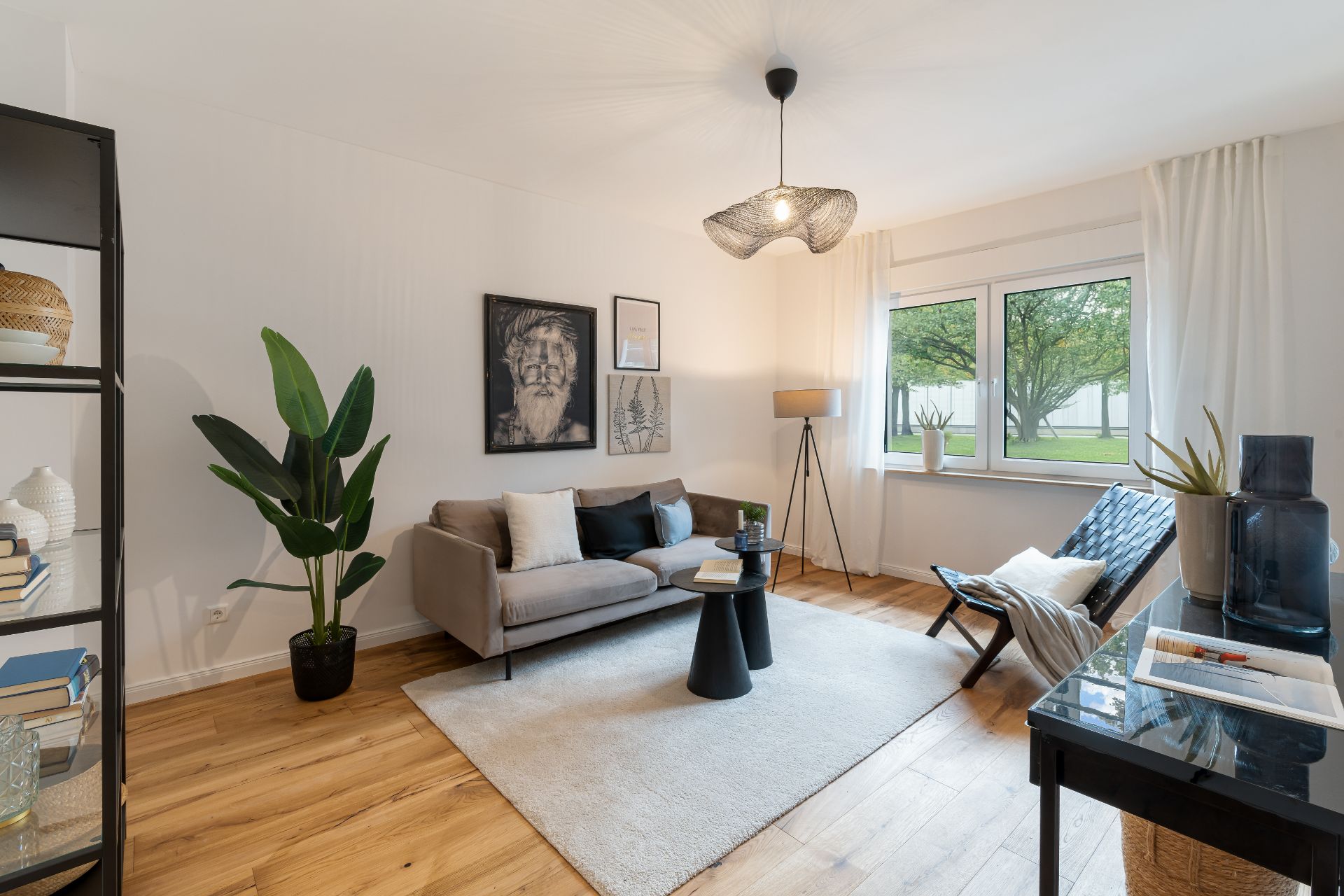 Helles Wohnzimmer mit Sofa, Sessel, Teppich, großen Fenstern und Zimmerpflanzen.