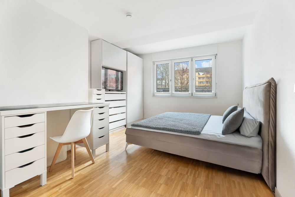 Helles Schlafzimmer mit Bett, Schreibtisch, Stuhl, Schrank und großem Fenster.
