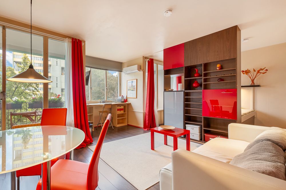 Helles Wohnzimmer mit roten Akzenten, großem Fenster, Esstisch, Sofa und Schreibtisch.