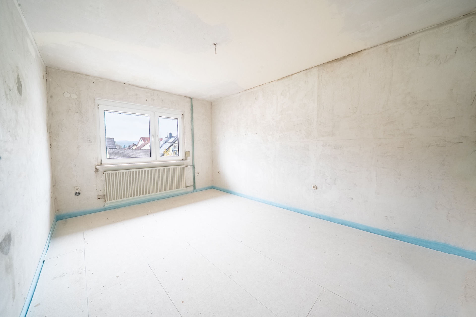Leerer, renovierungsbedürftiger Raum mit großem Fenster und Heizkörper, weiße Wände und Boden.