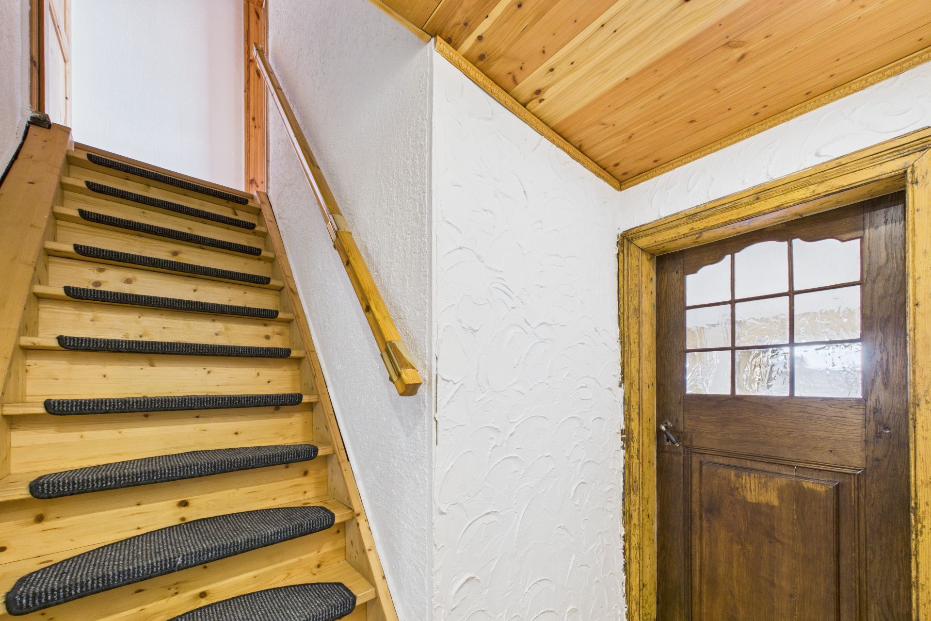 Hölzerne Treppe mit Handlauf, daneben eine braune Tür und Wand mit weißer Struktur.