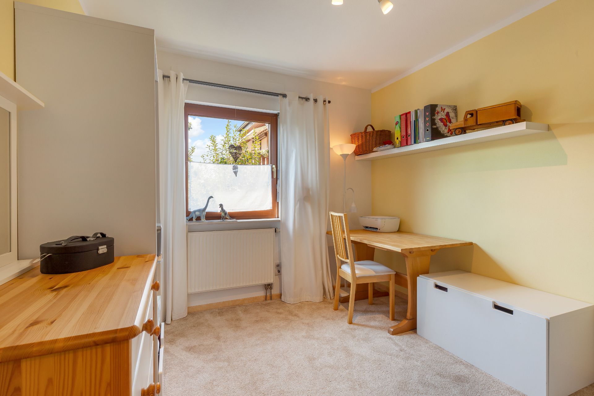 Helles Arbeitszimmer mit Holztisch, Stuhl, Regal und Fenster mit weißem Vorhang.