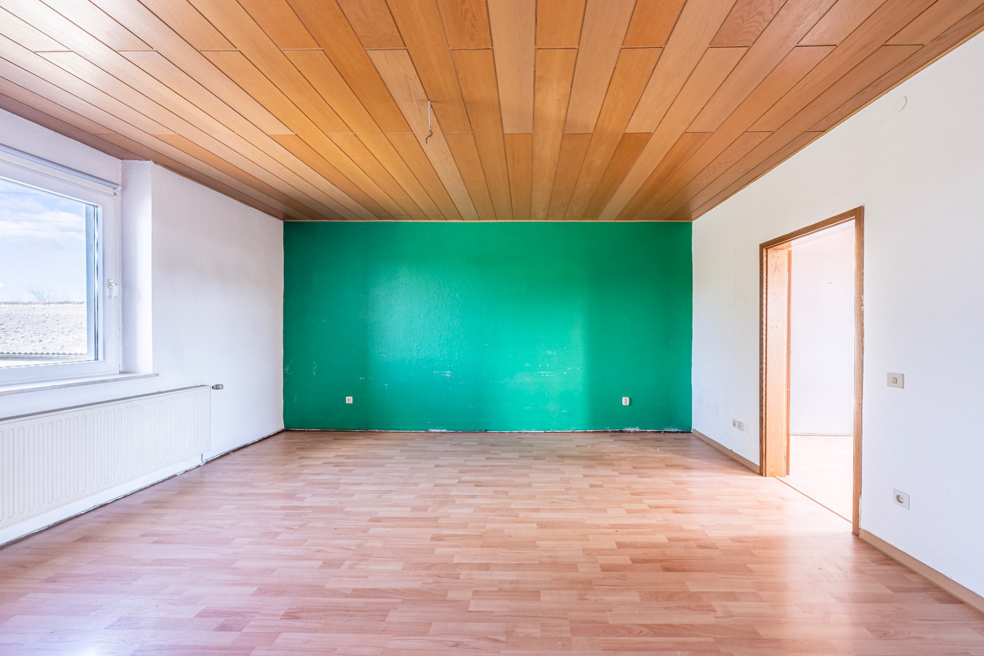 Leerer Raum mit Holzdecke, grüne Wand, Laminatboden und großem Fenster.