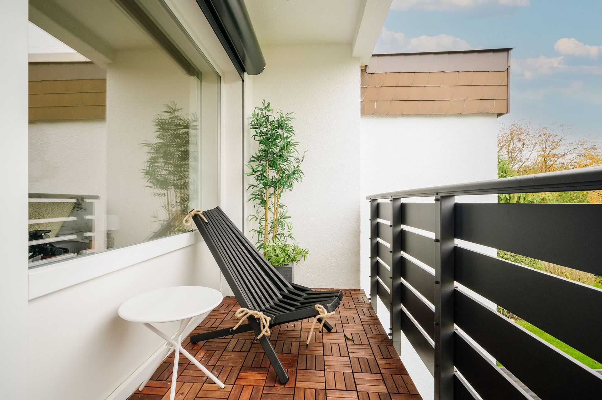 Balkon mit Holzfliesen, schwarzem Stuhl, weißem Tisch und grünem Pflanzkübel.